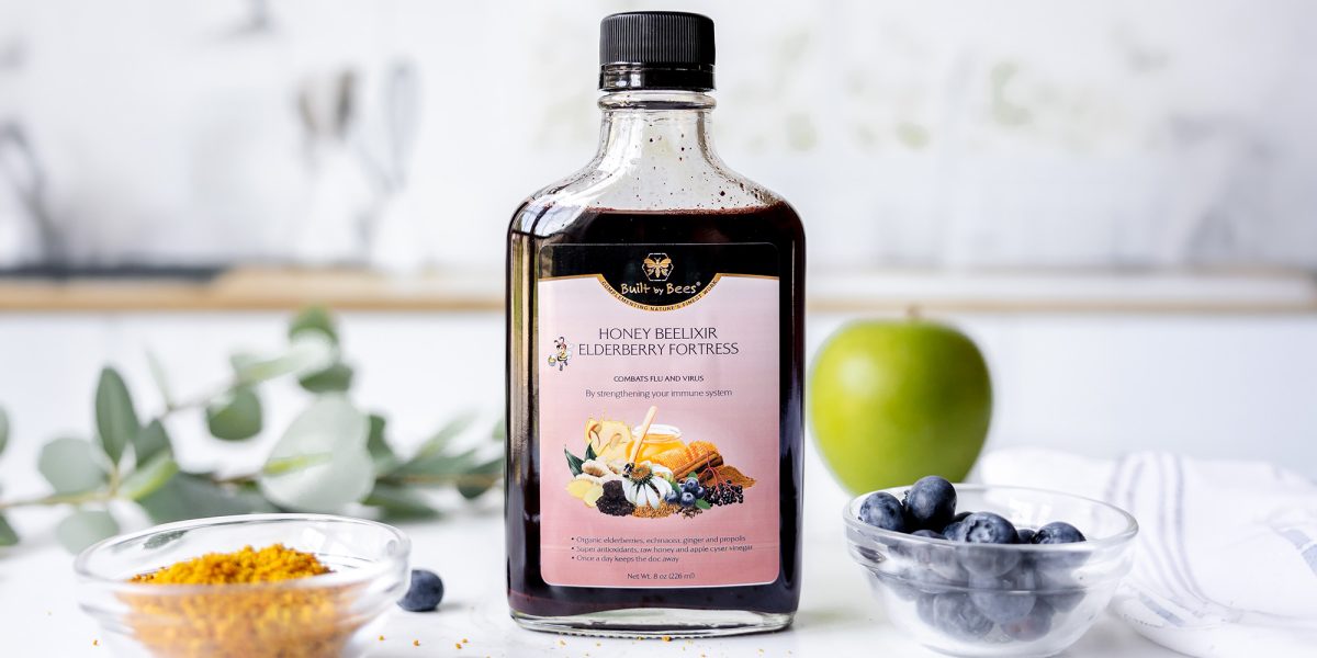 Bottle of 妻+无码+西瓜影音 honey elixir on countertop with bowl of bee pollen, fresh blueberries and a green apple