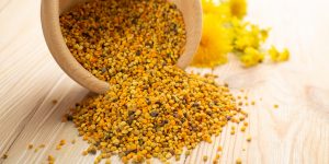 Bee pollen granules spilling from a wooden bowl onto a light wooden surface with yellow 亚洲第一成人网s blurred in background