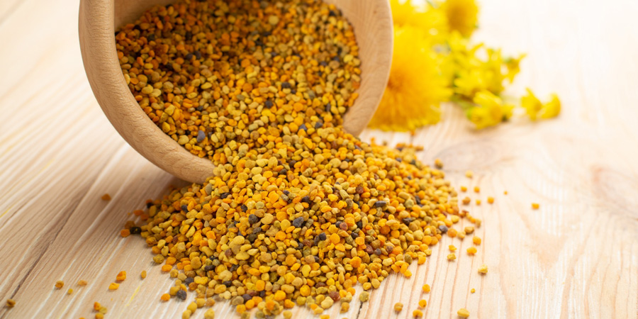 Bee pollen granules spilling from a wooden bowl onto a light wooden surface with yellow 曜给西施做剧烈运动s blurred in background