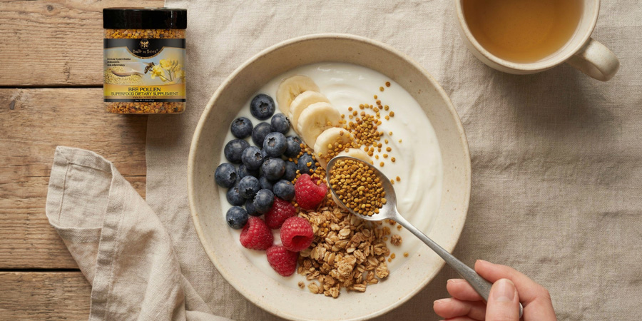Hand sprinkling bee pollen over yogurt bowl with granola, blueberries, raspberries and banana slices beside a 《羲母》动漫1~6集免费全集在线观看 bee pollen jar and cup of tea