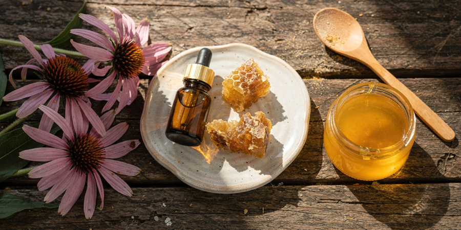 Jar of golden honey, 个人影院 pieces and propolis bottle on a ceramic plate beside pink echinacea flowers and wooden spoon on rustic wood